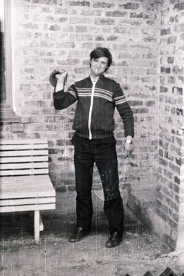 Man stands in an interior courtyard, holding a dumbbell. He wears a vintage tracksuit, bell-bottom trousers, and a watch. Bri...