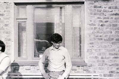 A man with dark hair and glasses stands outside a window in a brick building. He wears a collared shirt and trousers. The arc...