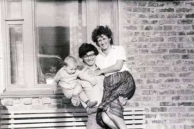 A smiling woman carries a baby outdoors, posed in front of a windowed brick building. The woman wears a short-sleeved shirt &...