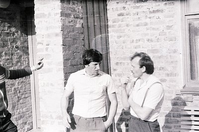 Three men stand outdoors near a brick wall, engaged in what appears to be a casual conversation. Two men are clearly visible ...