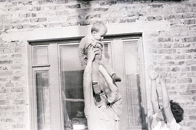A child is held aloft by an adult in front of a brick building with a multi-paned window. The image’s texture suggests a vint...