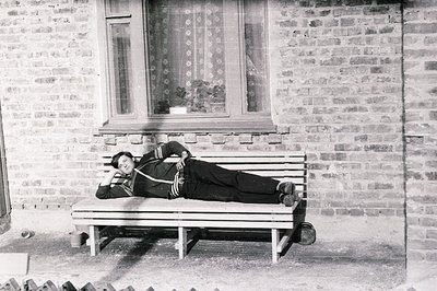A man reclines on a wooden bench outdoors, sporting a dark tracksuit and sneakers. Brick wall with a window featuring floral ...