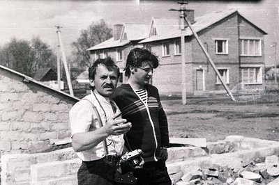 A man in workwear, suspenders, & a bricklayer, converses with a younger person in a striped sweater during what appears to be...