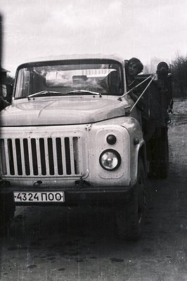 A utility truck, likely a Soviet-era UAZ-469, drives on a wet, gravel road. A man hangs from the door, smiling, while the dri...