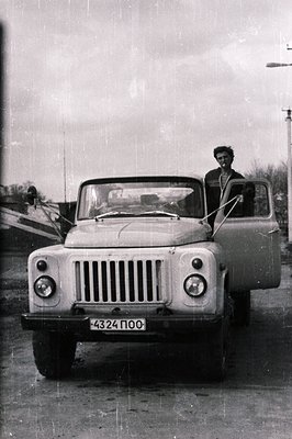A young man stands in an early model UAZ-69 jeep, door ajar, appearing to smile at the camera. The vehicle's Cyrillic license...