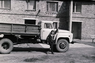 A young man stands leaning against a utilitarian Soviet-era truck, likely a UAZ-452, in front of a brick apartment building. ...