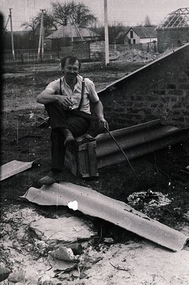 A construction worker, likely Bulgarian, sits on a wooden crate amidst debris and roofing materials. He wears a short-sleeved...