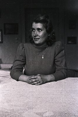 Vintage portrait of a woman seated on a bed, hands clasped. She wears a dark turtleneck sweater, a delicate necklace with a c...