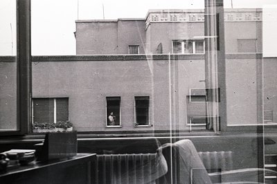 A monochrome image shows an exterior view through a window, likely an office or apartment. Visible is a modernist building wi...