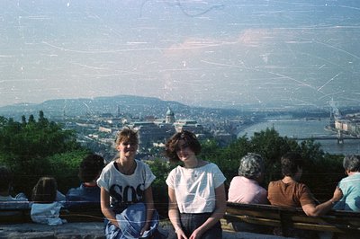 Two young people sit on tiered stone benches overlooking a cityscape, likely Budapest, Hungary. The panoramic view includes t...