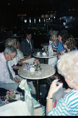 Group gathered at a table, likely a social gathering or formal event. Note 1970s/80s fashion and decor – men in suits, women ...