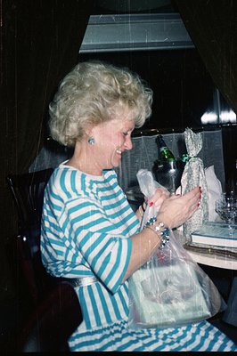A smiling woman, sporting a voluminous, blonde hairstyle, appears delighted while receiving a wrapped gift. She wears a blue ...