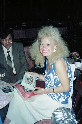A smiling woman with voluminous 1980s hair holds a wrapped gift and a book, seated at a restaurant table. A man in a suit sit...