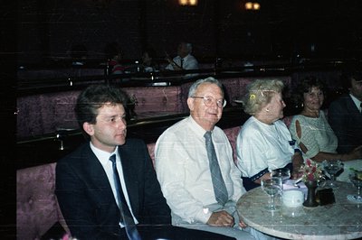 Three people seated on a plush, patterned red banquette. A young man in a suit sits beside an older gentleman in a button-dow...