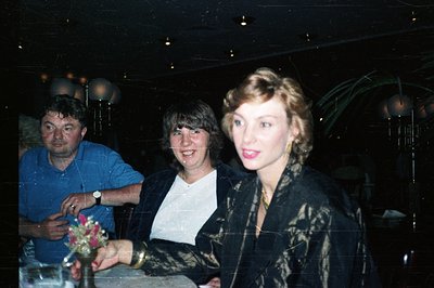 Three people stand in what appears to be a dimly lit, upscale event space. A man in a blue shirt stands to the left, two wome...