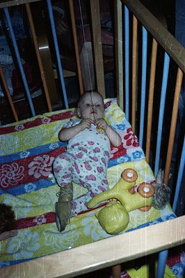 Infant in a crib, wearing patterned clothing, clutches a yellow plush toy. Crib bedding features a vibrant, geometric pattern...