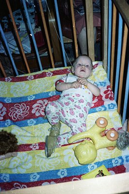 Infant seated in crib, wearing pink and white patterned clothing. Yellow rubber toy rests nearby. Appears to be a snapshot fr...