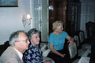 Three individuals seated in a formal living room, likely a family portrait. The man in a suit, two women in patterned blouses...