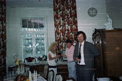 Formal dining room interior with three figures. A woman adjusts a vase, a man in slacks stands, and another in a suit poses. ...