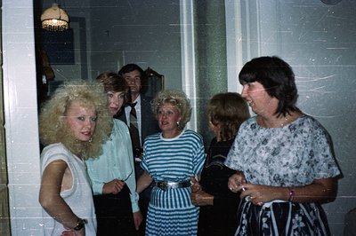 Candid moment: 5 people gather in a hallway, displaying 1970s fashion—large hair, wide collars, bold patterns. A man in a sui...