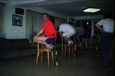 A staged scene featuring three men. One, wearing a red shirt and blue shorts, balances on a chair. Two other men are posed be...