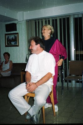 Man sits on a chair, smiling, with a woman in a pink tracksuit adjusting his collar. Interior scene, likely a recreation or s...