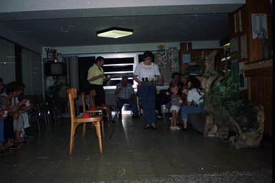Interior scene depicts a 1970s-era hotel lobby or function room. A server carries drinks past seated guests and a TV console....
