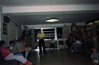 A group gathers in a sparsely furnished room, likely a recreational space, featuring a television, trophy display, and a wet ...