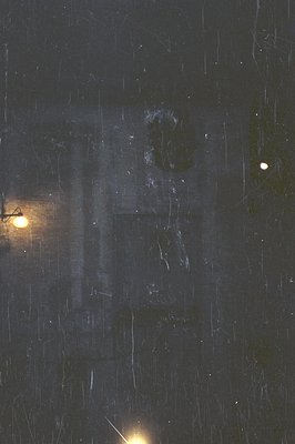 A blurred, rain-streaked exterior shot. A dimly lit doorway with a visible figure inside is partially obscured by water dropl...