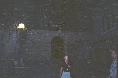 An exterior view featuring a woman and a man standing near a stone building with an arched doorway. The scene is dimly lit by...