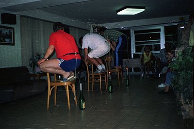 A group of people play "chair and bottle" in a dimly lit, modernist home. Three men, dressed in 1970s-era attire, balance on ...