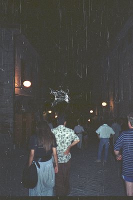 A group of people observes a suspended, ornate chandelier within a dark, arching passageway. The building features stone wall...