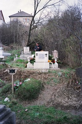 An elderly man stands before a memorial in a hillside cemetery, possibly Eastern Europe. Stone monuments and graves are visib...