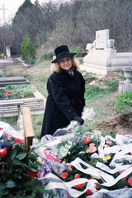 A woman in a black coat and hat tends to a memorial covered with flowers and ribbons. The grave site features a large, ornate...