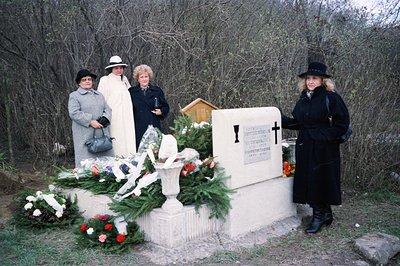 Four women in formal dress stand before a marble monument adorned with floral arrangements, possibly at a memorial or gravesi...