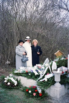Three women stand before a stone monument heavily adorned with floral tributes. The women wear 1970s-era clothing and hats. A...