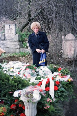 A woman in a dark coat stands before a lush floral arrangement at a gravesite. An inscription on a nearby headstone is visibl...