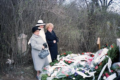 Three women in vintage attire stand beside a large arrangement of fresh flowers, likely at a gravesite or memorial. The women...