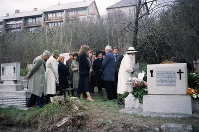 Group gathers at a gravesite in a European cemetery. The scene features floral tributes, a stone cross, and a memorial plaque...