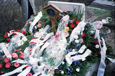 Elaborate floral arrangement with chrysanthemums, lilies, and ribbons adorns a gravestone. A small, wooden plaque is prominen...