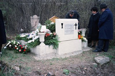 A solemn scene at a gravesite, featuring a large, ornate stone memorial adorned with fresh flower arrangements and a small, c...