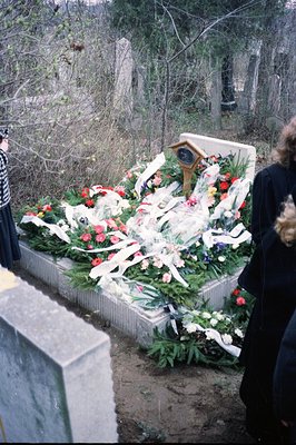 Overgrown concrete memorial platform, heavily adorned with floral tributes and ribbons. The platform appears to be within a w...