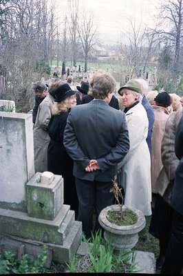 A group gathers at a weathered grave site, likely during a memorial or remembrance. Stone monuments line the hillside. Indivi...
