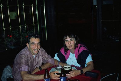 Two individuals sit at a table with drinks during nighttime; a beer and bottled beverages visible. The man wears a patterned ...