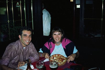 Two people seated at a table with a pizza, likely outdoors at night. Man in a short-sleeve shirt smiles directly at the camer...