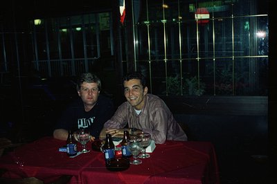 Two men seated at a table covered with a vibrant red tablecloth, appear to be enjoying drinks, likely beer. The setting is ou...