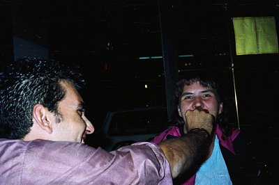 Two men share a playful moment in a dimly lit, possibly outdoor setting. One man, in a purple shirt, smiles while appearing t...