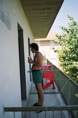 A man stands on a balcony, holding a can, appearing relaxed in patterned swim trunks and flip-flops. Laundry hangs to dry on ...