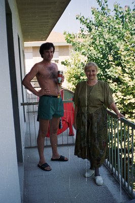 A man and older woman stand on a balcony; the man is shirtless, wearing swim trunks & sandals, holding a drink. The woman wea...