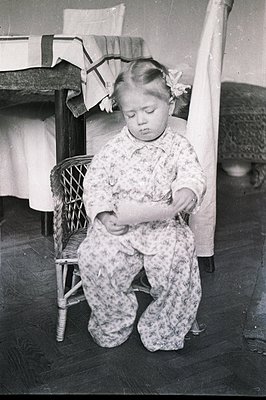 A young girl, likely toddler age, kneels on a wood floor before a rattan chair. She wears a patterned floral dress and a flow...